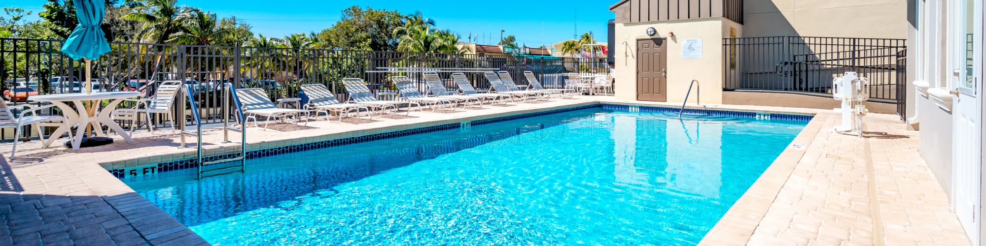 A clear, blue outdoor swimming pool with lounge chairs and tables with umbrellas around it, under a bright sky, next to a building.