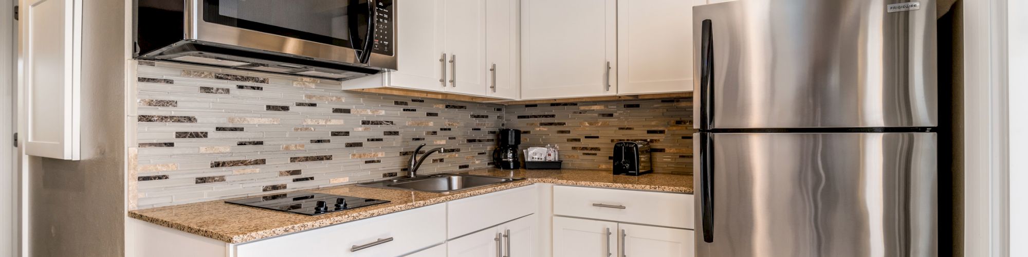 A modern kitchen with stainless steel appliances, including a refrigerator and microwave, white cabinets, tiled backsplash, and granite countertops.