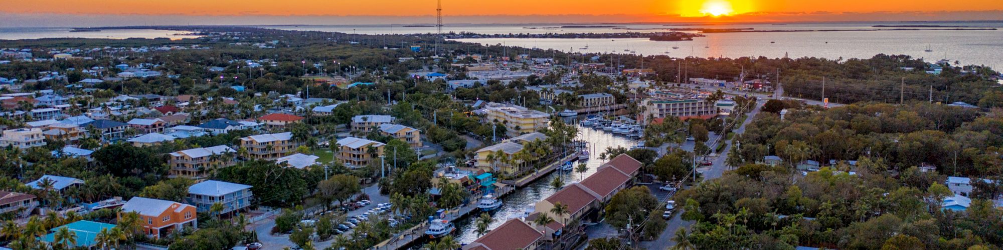 An aerial view of a coastal town at sunset, with houses, boats, and waterways in the foreground and the sun setting over the horizon.