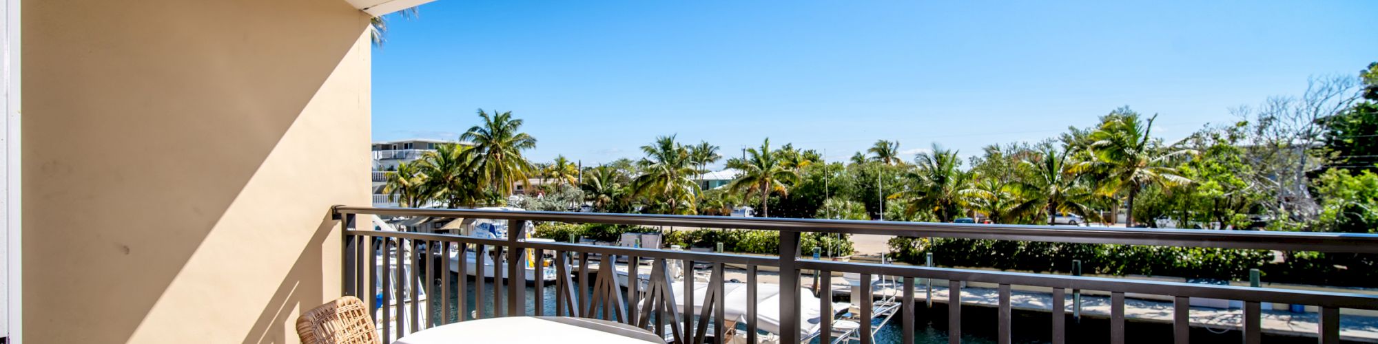 A balcony with a small table and two chairs, overlooking a marina with boats and palm trees, under a clear blue sky.