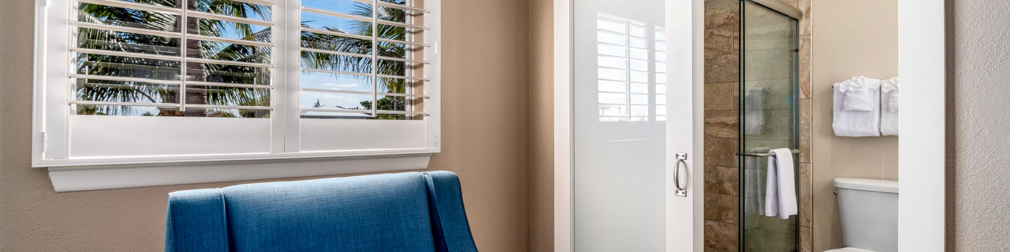 A small room with a blue chair, window with blinds, and adjacent bathroom featuring a sliding door, toilet, and shower.