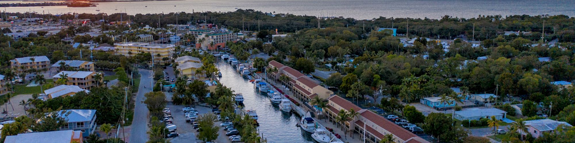 Aerial view of a coastal town at sunset, featuring a waterway lined with boats, surrounding buildings, trees, and a glowing horizon over the ocean.