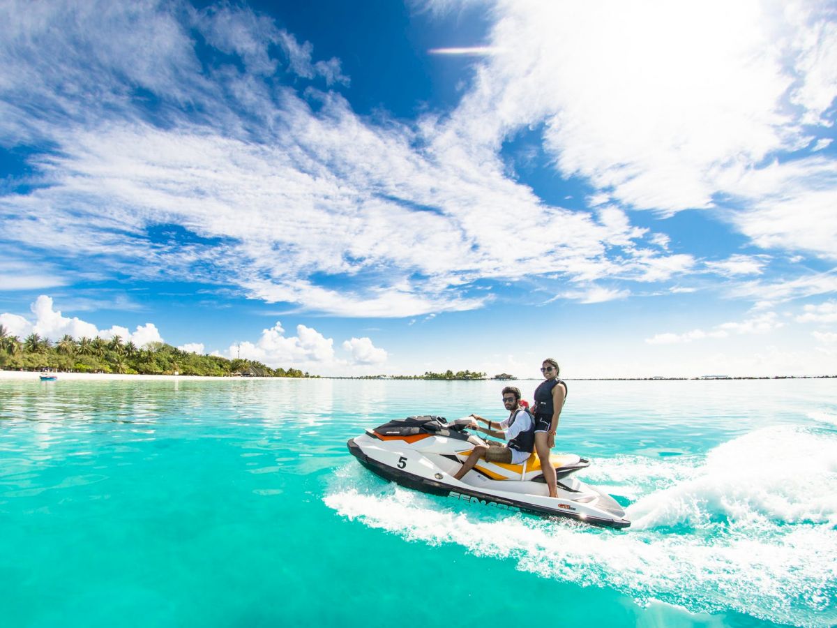 Two people are riding a jet ski on clear turquoise water with a beautiful sky and some distant trees on a sunny day.