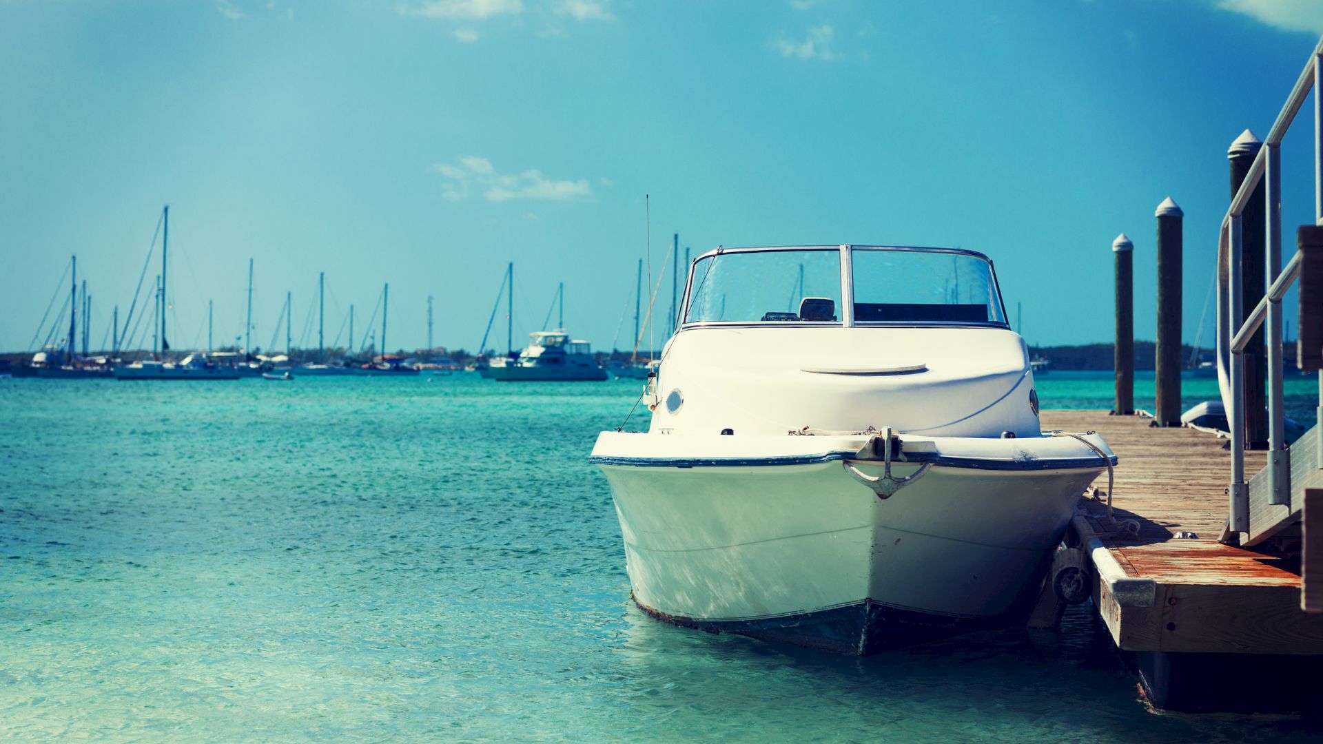 A white motorboat is docked at a wooden pier in clear blue waters with multiple sailboats in the background under a clear sky.
