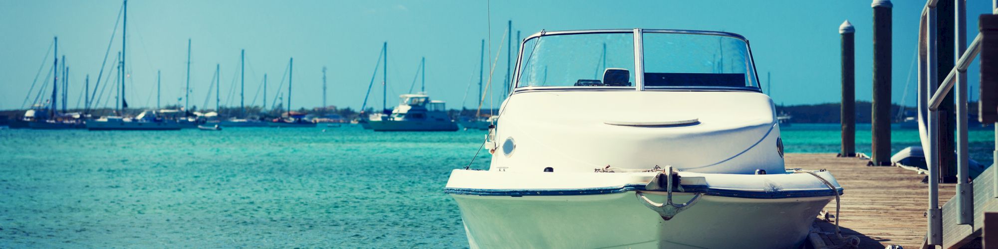 A white motorboat is docked at a wooden pier in clear blue waters with multiple sailboats in the background under a clear sky.