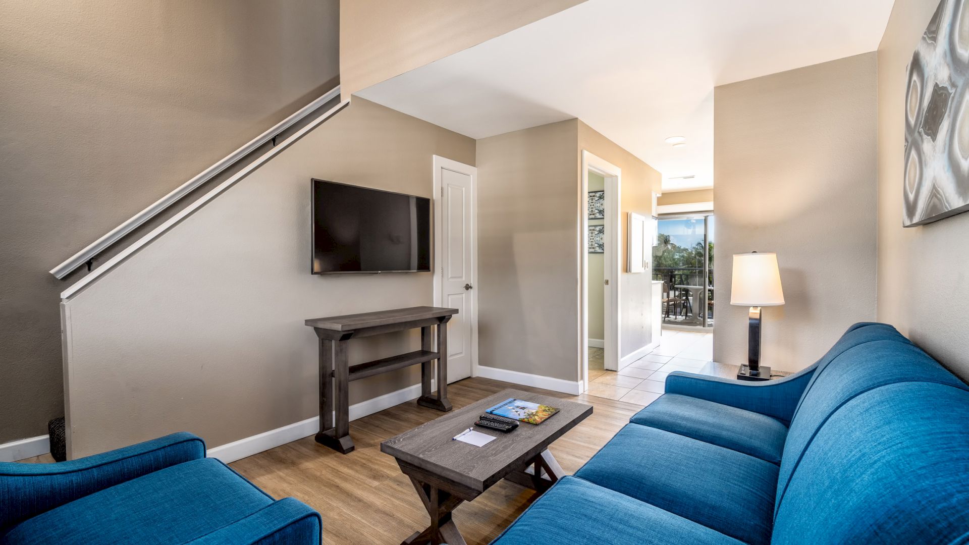 A cozy living room with blue furniture, a wall-mounted TV, a coffee table, a lamp, stairs, and a hallway leading to another room ends the sentence.