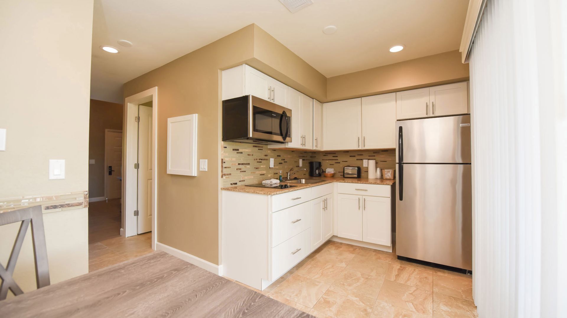 Modern kitchen with white cabinets, stainless steel appliances, and beige walls. It includes a microwave, refrigerator, and a light wood dining table.