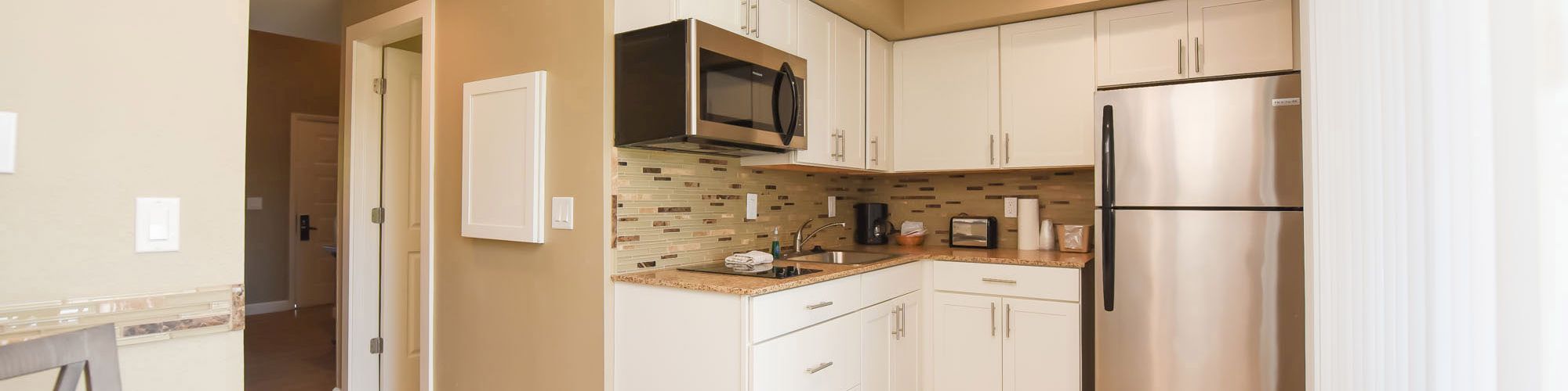 The image shows a modern kitchen with white cabinets, stainless steel appliances, and beige walls. It includes a microwave, refrigerator, and a light wood dining table.