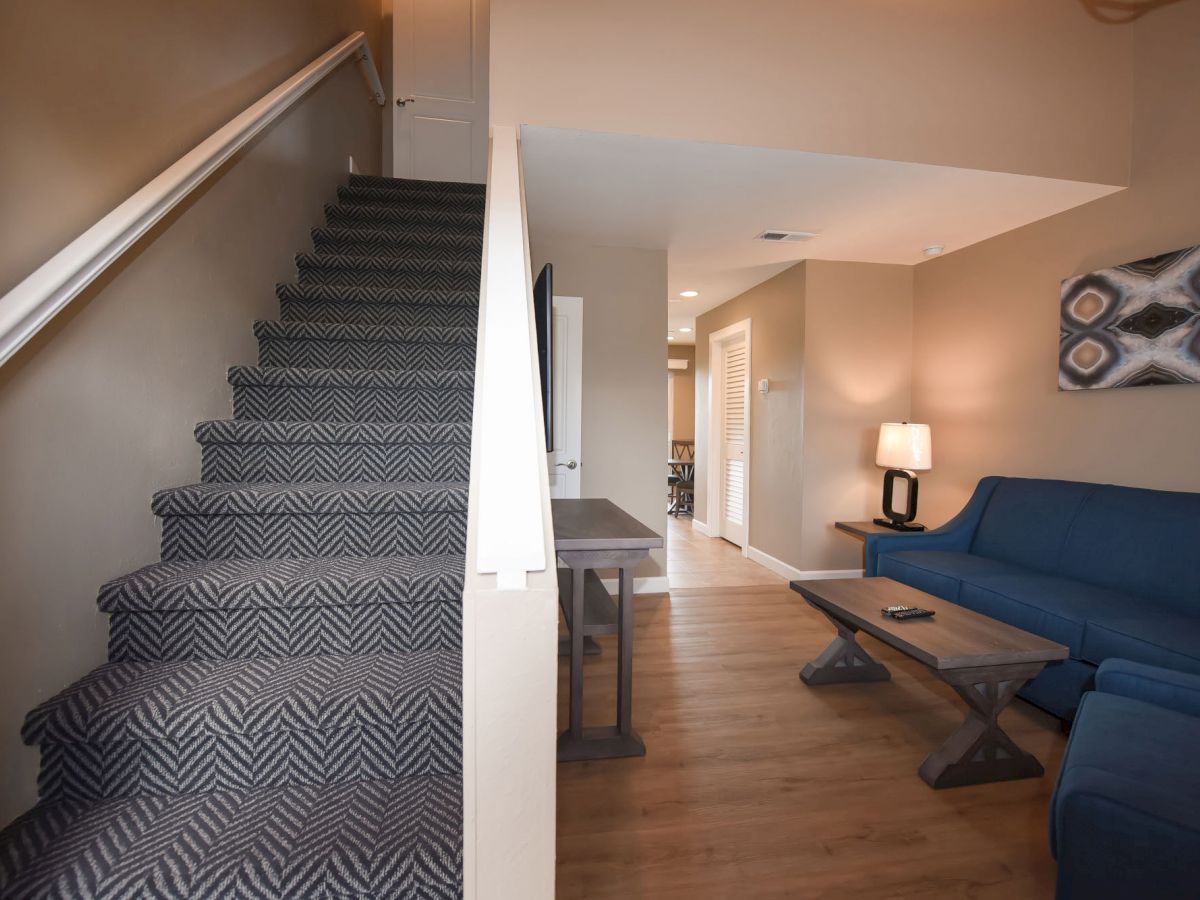 A living area with blue furniture, wooden flooring, a staircase with patterned carpet, a table lamp, a wall art piece, and a hallway.