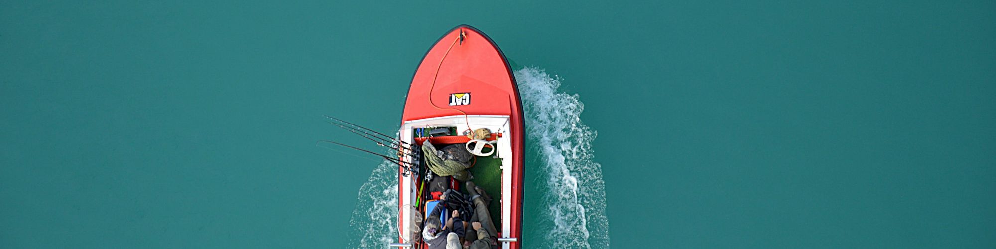 A red boat is moving across turquoise water, carrying two individuals and some gear, leaving a wake behind it.