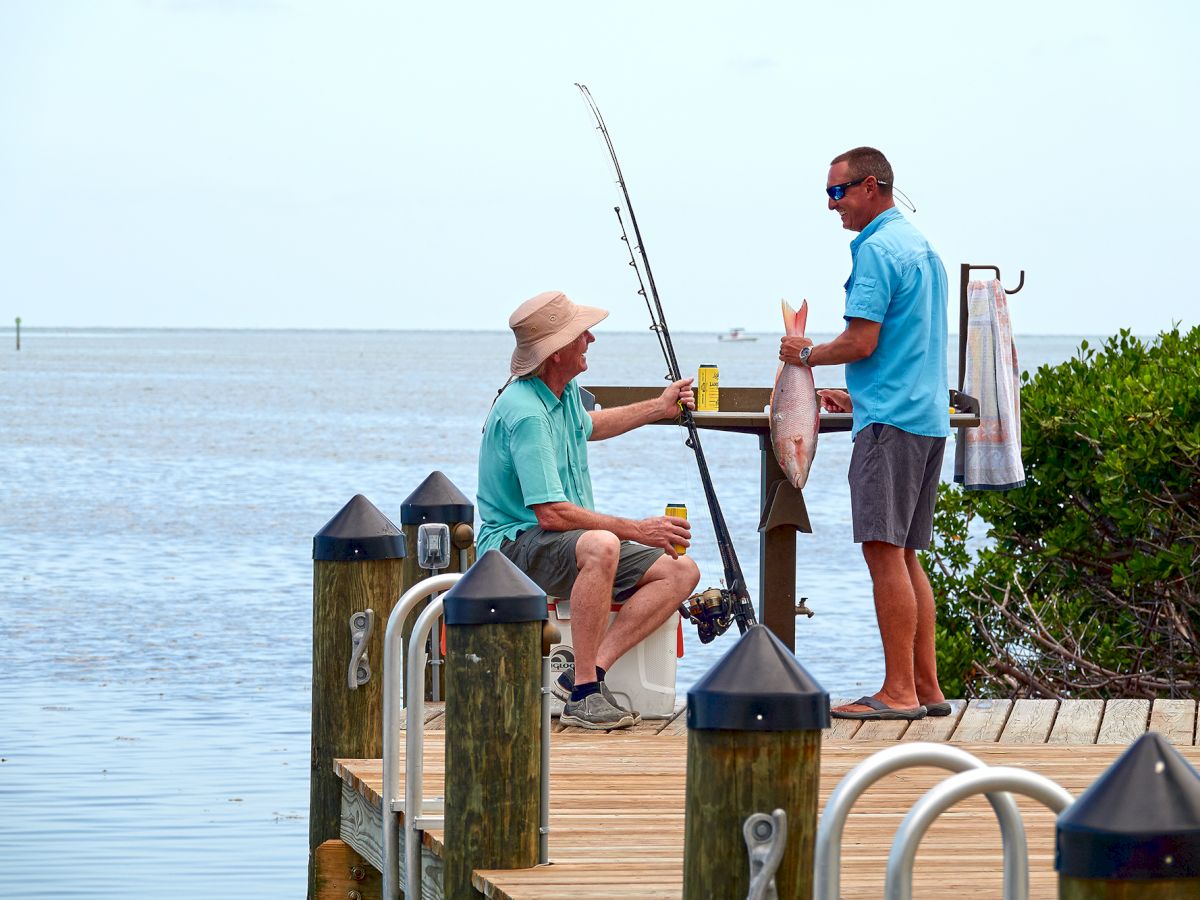 Two people are on a pier by the water; one is sitting with a fishing rod and a drink, and the other is standing, holding a fish and a drink.