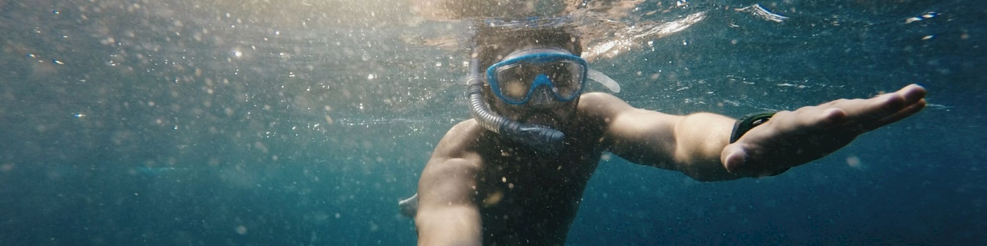 A person is underwater, wearing a snorkeling mask and extending an arm forward, possibly taking a selfie. Sunlight filters through the water.