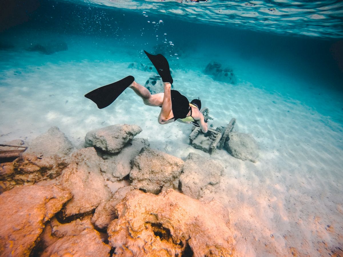 A person is snorkeling underwater, exploring rocks and marine life, wearing flippers and a mask in clear, blue water.