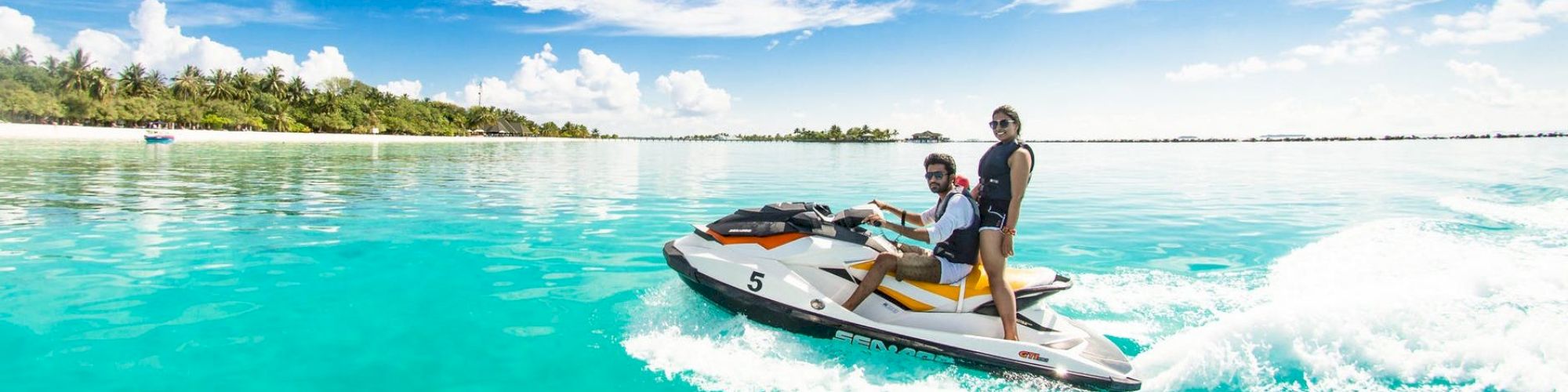 Two people riding a jet ski on clear turquoise water near a tropical island with a blue sky and fluffy clouds.