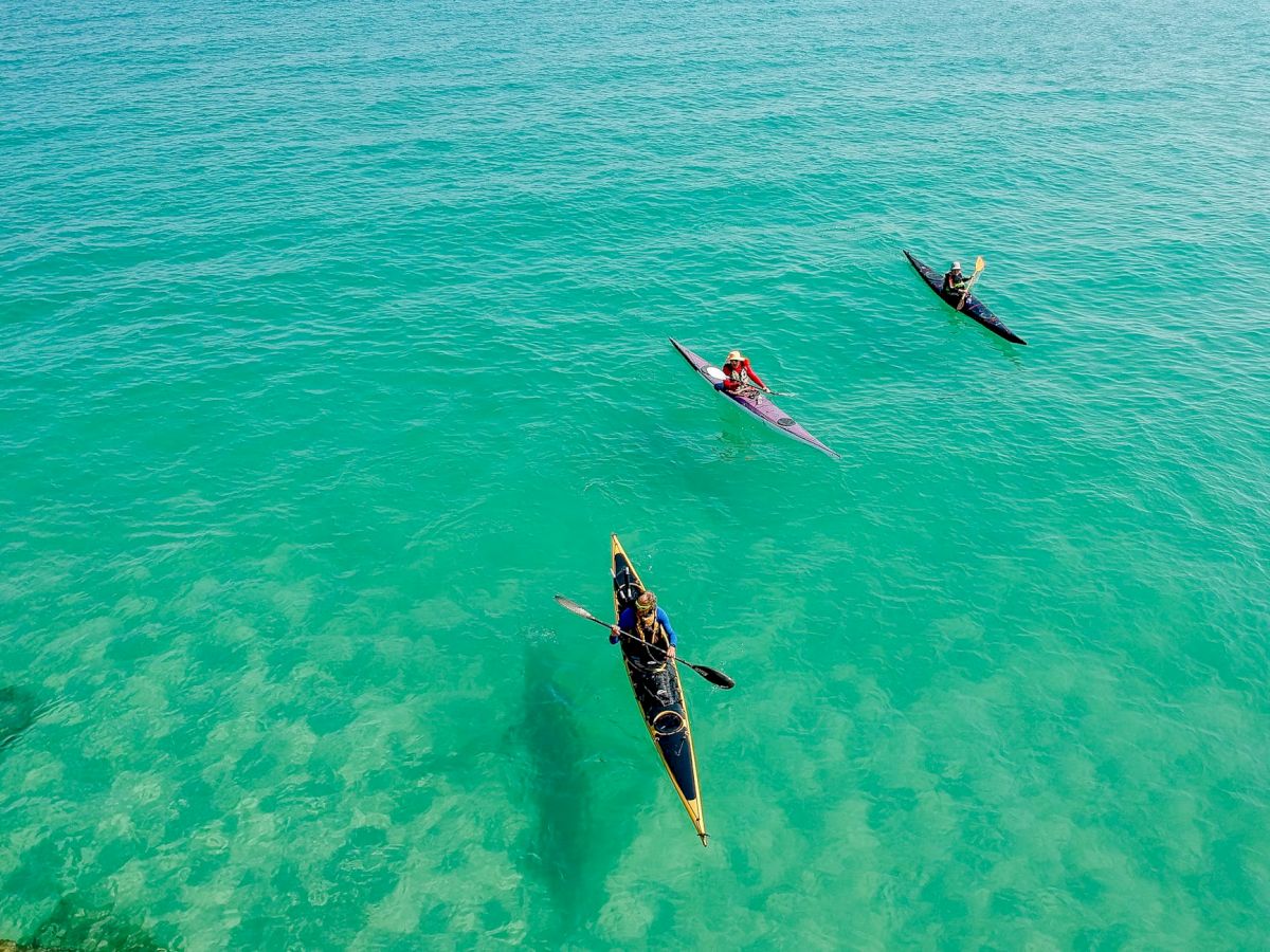 Three kayakers paddle in clear turquoise water, with sunlight highlighting the ocean floor below.