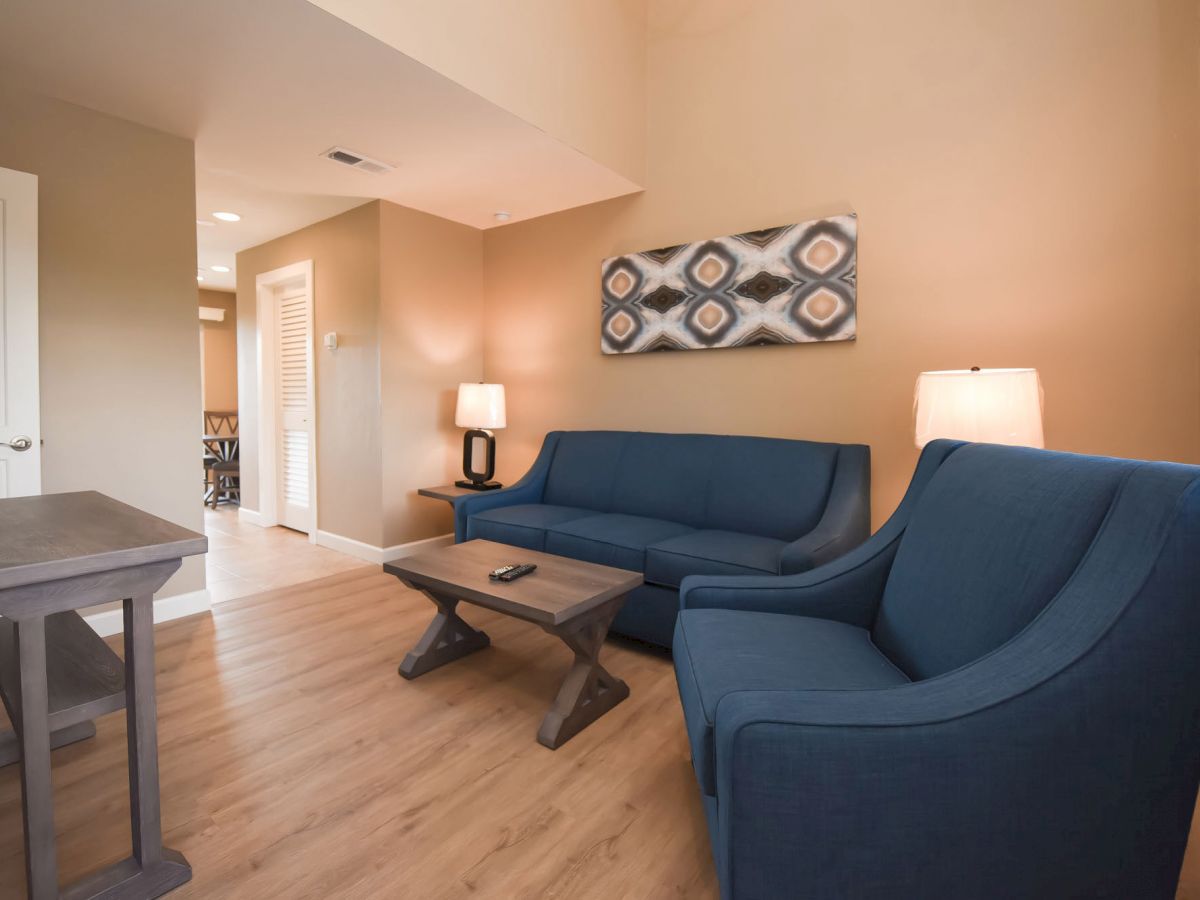 A cozy living room with blue sofas, wooden furniture, patterned wall art, and warm lighting, featuring two lamps and a remote on the coffee table.