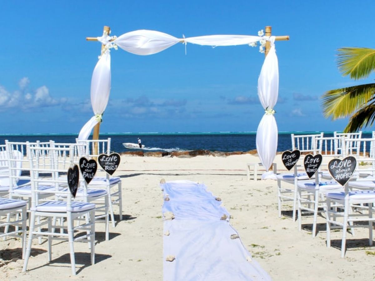 A beachside wedding setup with white chairs, heart decorations, a white aisle runner, and an arch overlooking the ocean under a sunny sky.