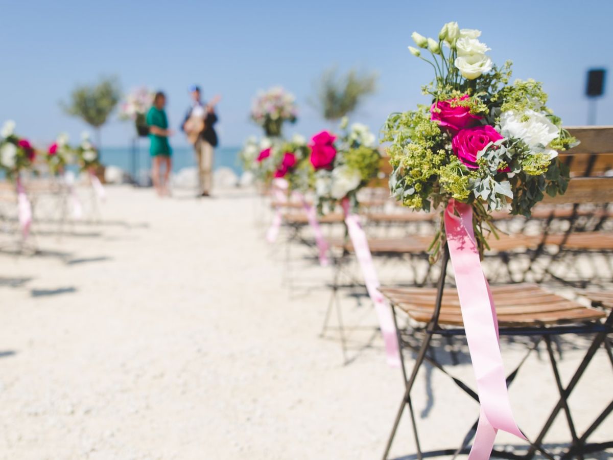 A beachside outdoor wedding setup with chairs decorated with flowers and pink ribbons, people in the background, and a clear blue sky.