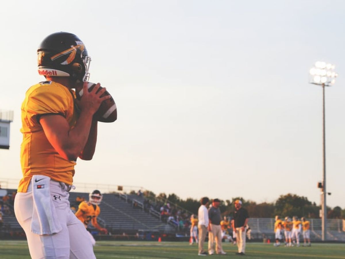 A football player in a yellow jersey holds a ball, preparing to throw during a practice session on a field at sunset.