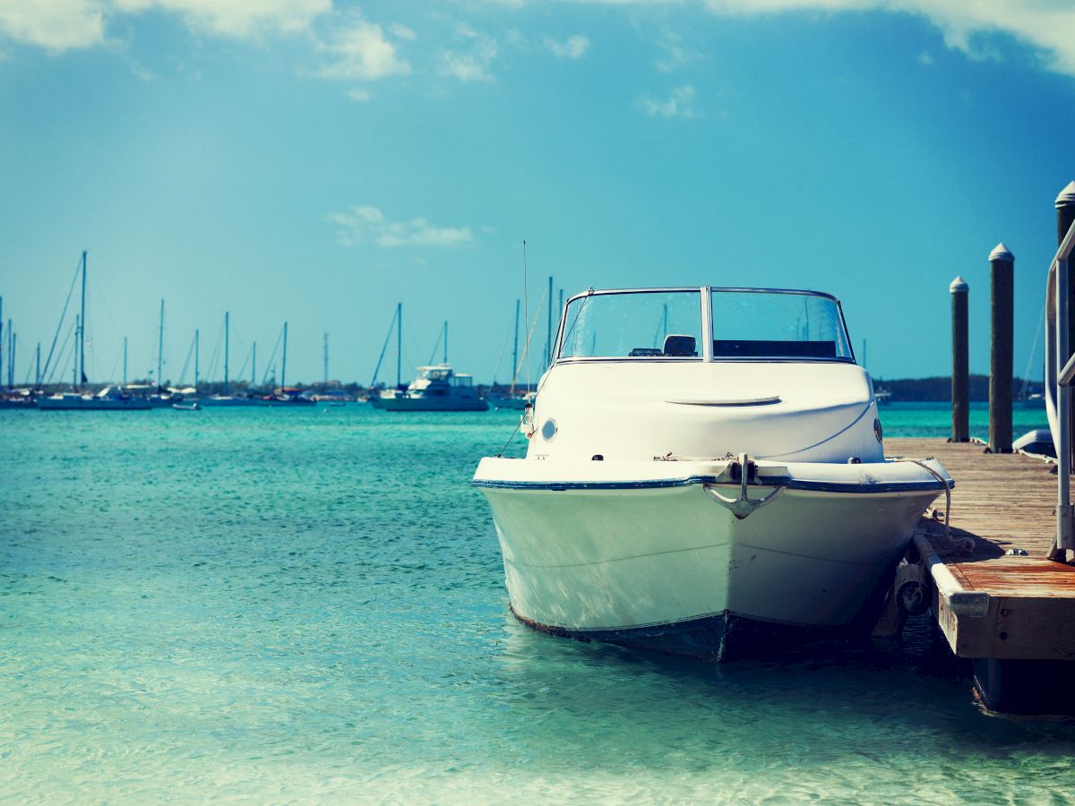 A small white boat is docked at a pier against a backdrop of a turquoise sea with several sailboats anchored in the background under a clear sky.