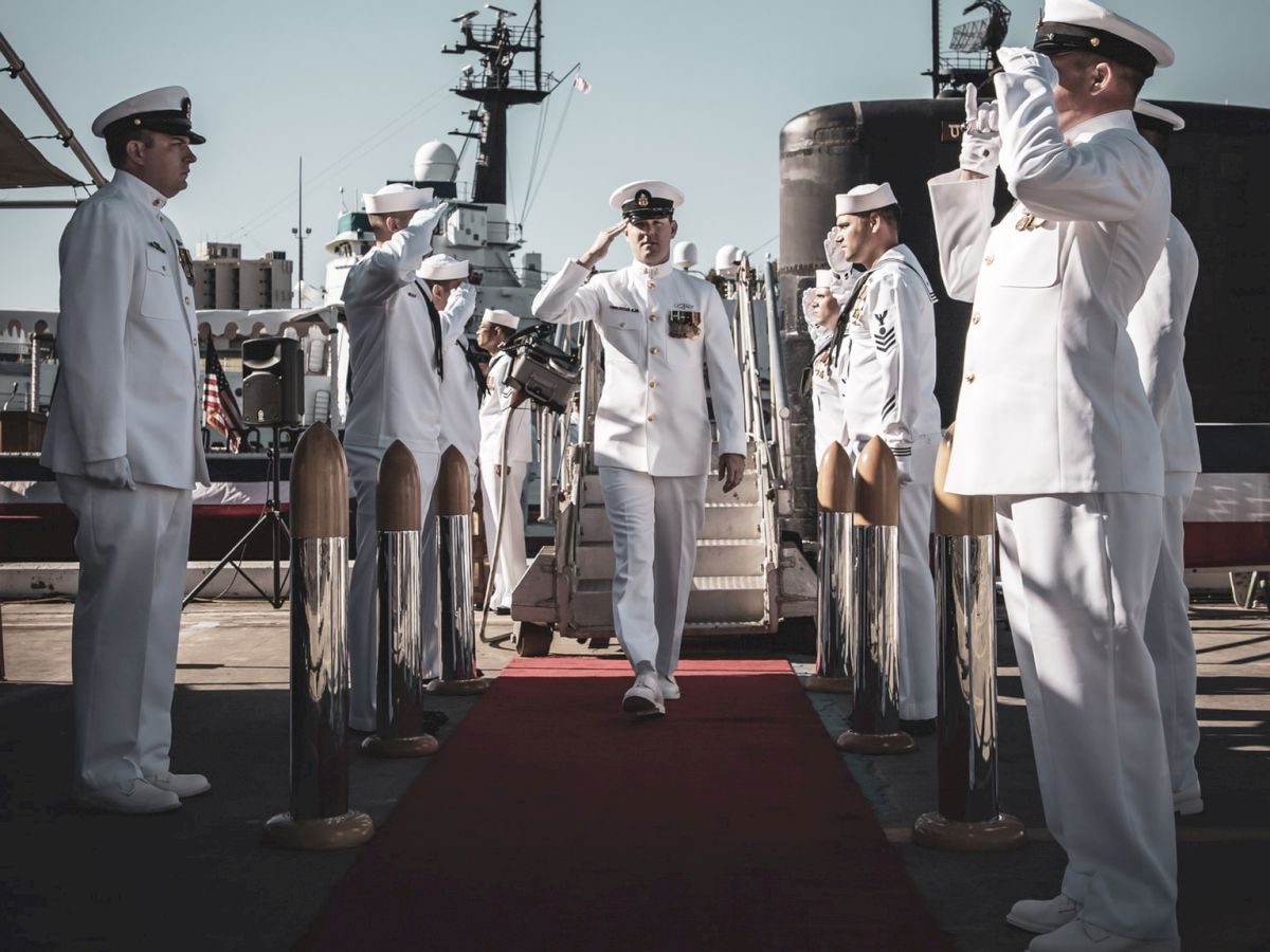 A naval officer walks on a red carpet, saluting as others in white uniforms stand on either side, also saluting.