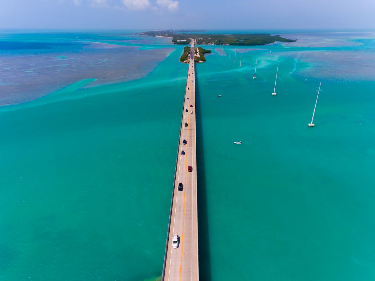 The image shows a long bridge stretching across vibrant turquoise waters, connecting two landmasses under a clear sky with a few clouds.