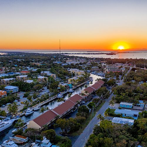 An aerial view of a coastal town at sunset, showing buildings, boats, and the sea in the distance with an orange sky.