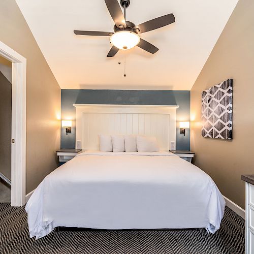 A bedroom with a made bed, ceiling fan, artwork on the wall, nightstands with lamps, and a doorway leading to another room or hallway.