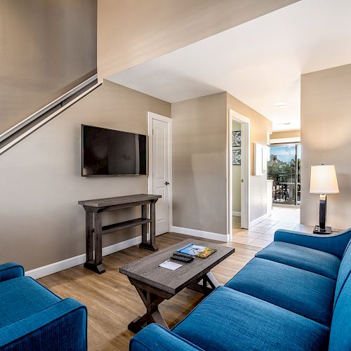 A cozy living room with blue seating, a wall-mounted TV, a coffee table, and a lamp. Stairs lead up while a door opens to another space.