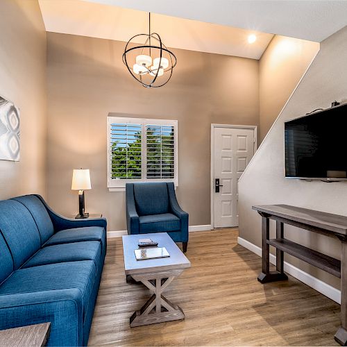 The image shows a modern living room with a blue sofa, armchair, coffee table, flat-screen TV, wall art, and a window featuring white blinds.