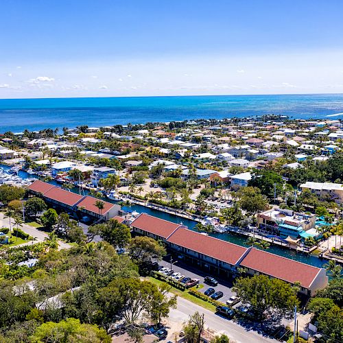 A coastal town with numerous houses, tree-lined streets, and a waterway leading to the ocean under a clear blue sky.