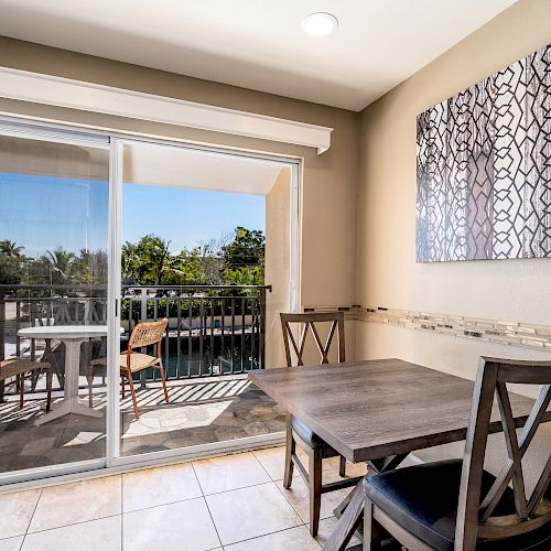 The image shows a dining area with a wooden table and two chairs, a sliding glass door to a balcony, and abstract wall art above the table.