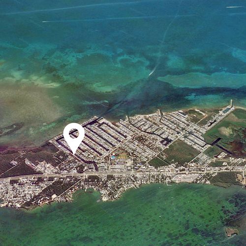 Aerial view of a coastal town with clear blue water, outlined land and a map pin marking a specific location within the town.