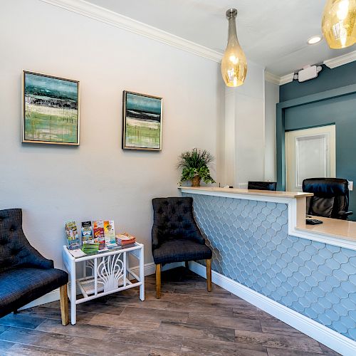 This image shows a modern office reception area with two blue chairs, a small table with magazines, a reception desk, and wall art.