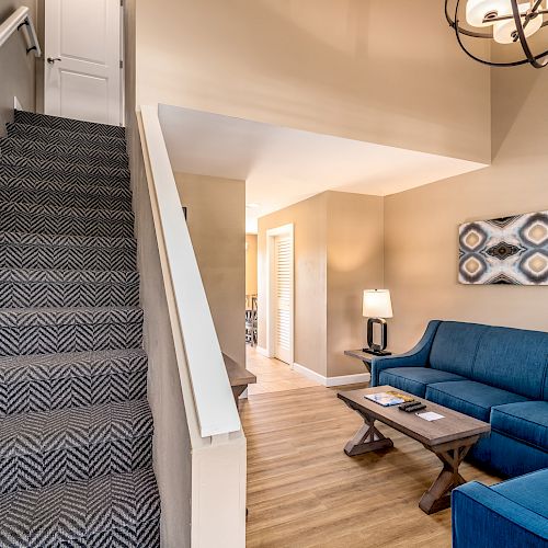 The image shows an interior space with a staircase on the left, a blue sofa set, a coffee table, and a wall-mounted artwork on the right side.