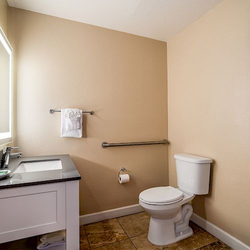 A bathroom with a toilet, sink, wall-mounted towel rack, two framed pictures, and a lit mirror above the sink, featuring beige walls and tile flooring.