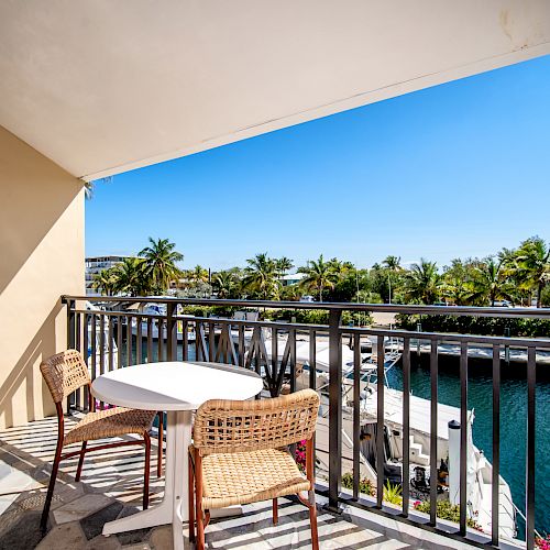 A balcony scene with a table and chairs overlooks a serene waterfront, lined with palm trees under a bright blue sky, providing a tranquil view.