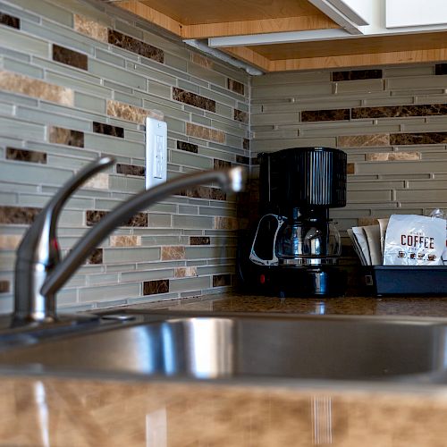 The image shows a kitchen sink with a faucet, a tile backsplash, and a coffee maker with coffee supplies placed on the countertop.