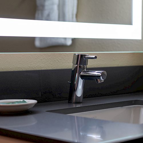 A modern bathroom sink with a chrome faucet, an illuminated mirror, and a dish on the counter containing soap, reflecting a clean, contemporary design.