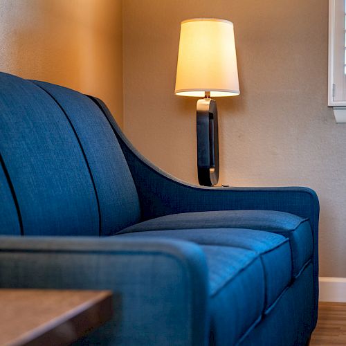 A cozy living area with a blue upholstered couch, a side table, a modern table lamp, and a window with white shutters can be seen.