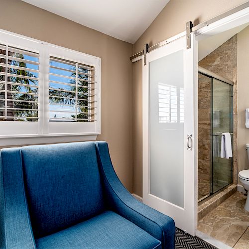 A bathroom with a sliding door, a toilet, a glass shower, and a blue chair near a window with white shutters is shown in the image.