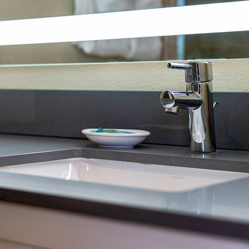 A modern bathroom sink with a sleek faucet, a small dish holding a few items, and a well-lit mirror in the background.