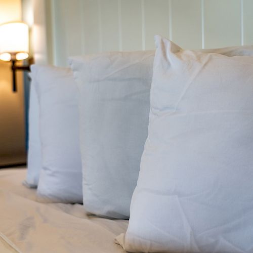 The image shows a neatly made bed with three white pillows lined up, and a wall-mounted lamp is visible in the background.