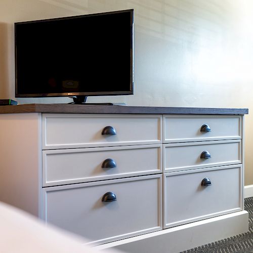 A modern TV sits on top of a white dresser with six drawers, against a beige wall in a room with carpeted flooring.