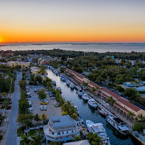 Aerial view of a coastal town at sunset with a canal lined with boats and surrounded by greenery and buildings. The sun sets over the horizon.