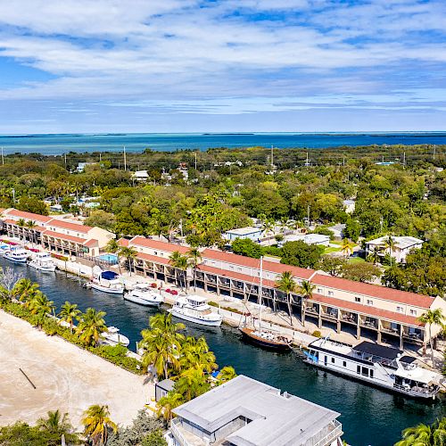 Aerial view of a waterfront community with boats docked along a canal, surrounded by greenery, buildings, and a distant blue sea.