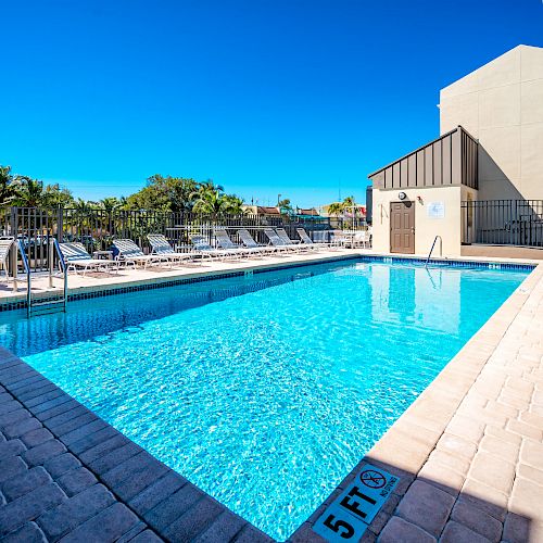 An outdoor swimming pool with clear blue water is surrounded by lounge chairs, umbrellas, and a building nearby under a clear, sunny sky.