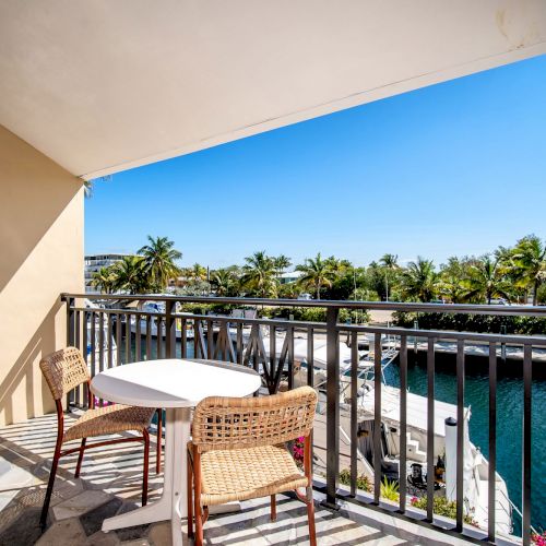 A balcony with two chairs and a table overlooks a canal lined with palm trees under a clear blue sky.