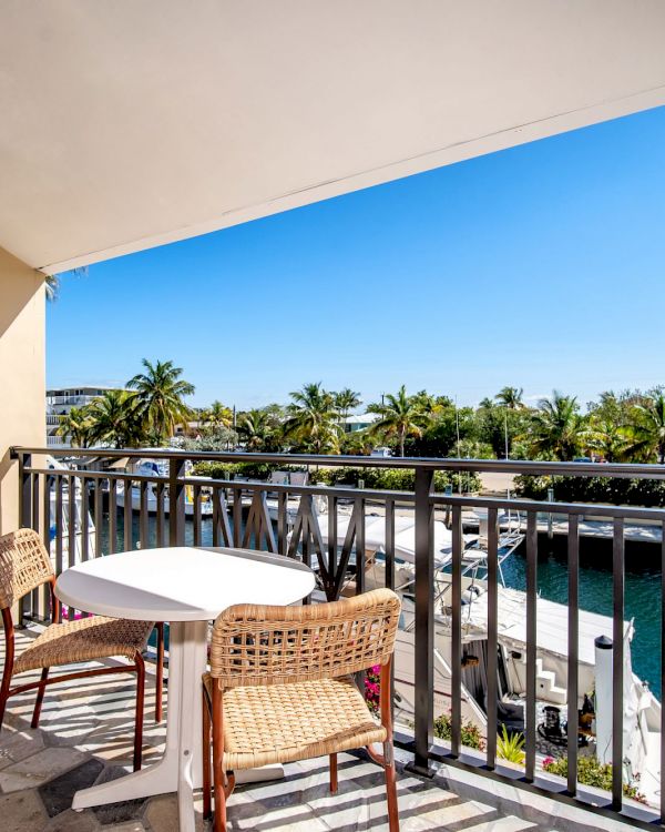 A balcony with two chairs and a table overlooks a canal lined with palm trees under a clear blue sky.