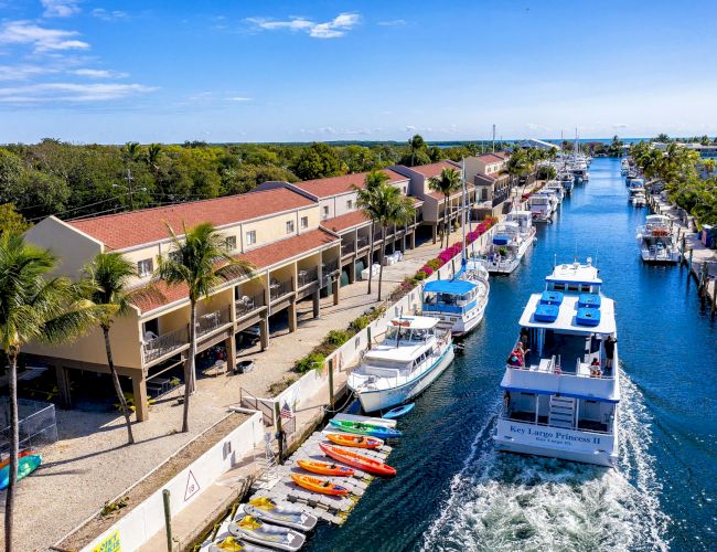 A canal with boats, kayaks, and waterfront buildings surrounded by greenery under a blue sky, creating a picturesque and serene scene.